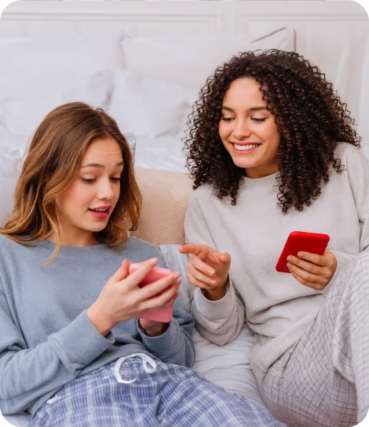 Two friends sitting on a bed, smiling while looking at their phones
